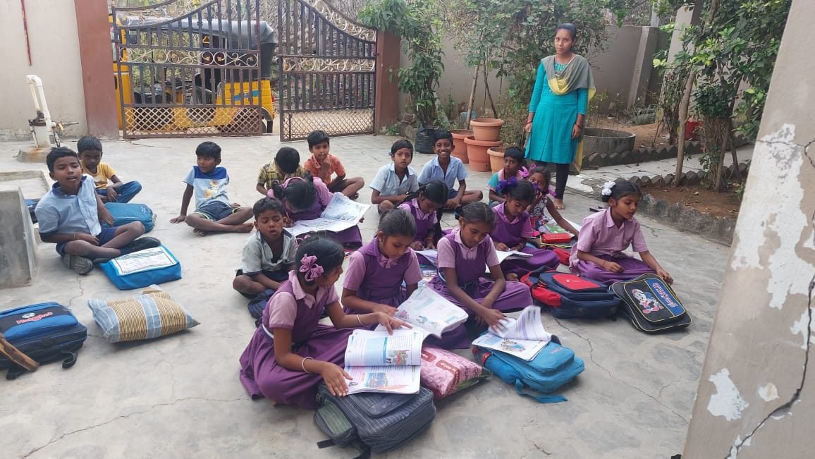 Children seated outdoors with open textbooks and school bags, a teacher standing nearby